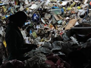 Descrição da imagem: Silhueta de uma mulher em frente a uma pilha de lixo reciclavel que preenche toda a imagem. Ela separa os reciclaveis.